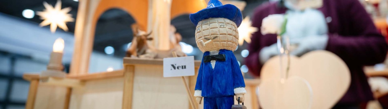 Im Vordergrund ist das geschnitzte Leipziger Messemännchen zu sehen, im Hintergrund sind – leicht verblasst – weitere Holzfiguren sowie die Ausstellerin auf der HANDWERK live zu erkennen.