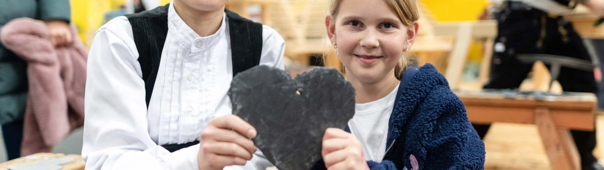 Eine Auszubildende der Dachdeckerinnung hält auf der HANDWERK Live gemeinsam mit einem Mädchen ein zusammengeschiefertes Herz hoch.
