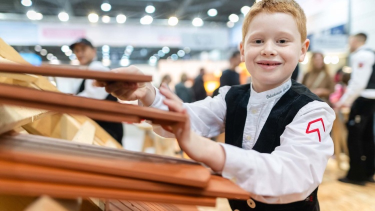 Ein kleiner Junge in Dachdeckerkleidung strahlt auf der HANDWERK live in die Kamera, während er Dachziegel in den Händen hält.