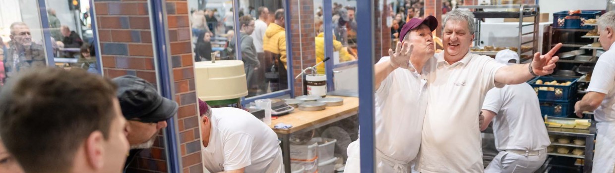 Mit lustigen Grimassen winken zwei Bäcker hinter einer Glasscheibe in die Kamera, während Besuchende der HANDWERK live die Gläserne Bäckerei bestaunen.