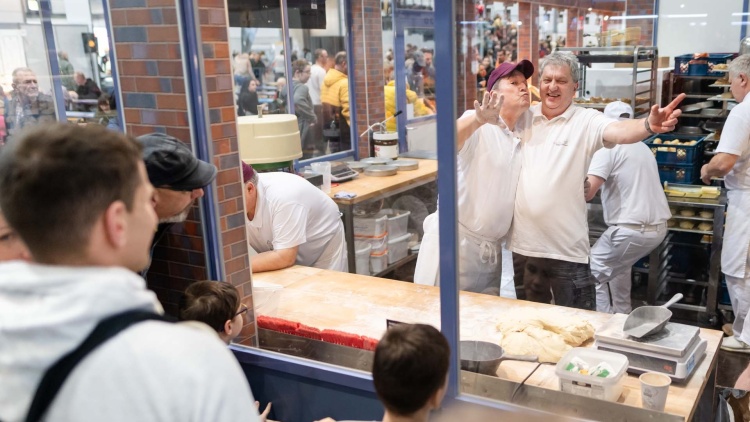 Mit lustigen Grimassen winken zwei Bäcker hinter einer Glasscheibe in die Kamera, während Besuchende der HANDWERK live die Gläserne Bäckerei bestaunen.