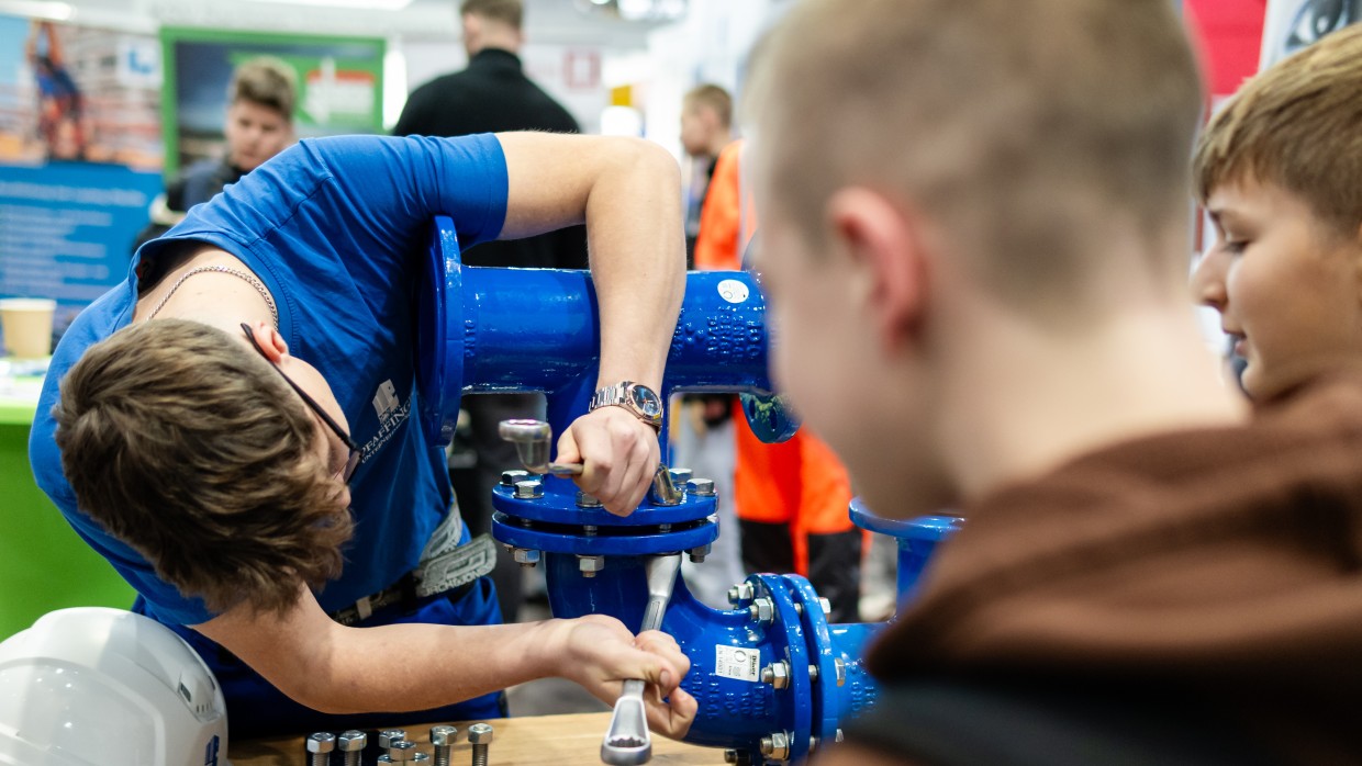 Ein Handwerker zeigt Kinder das befestigen von Rohren.