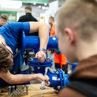 Ein Handwerker zeigt Kinder das befestigen von Rohren.