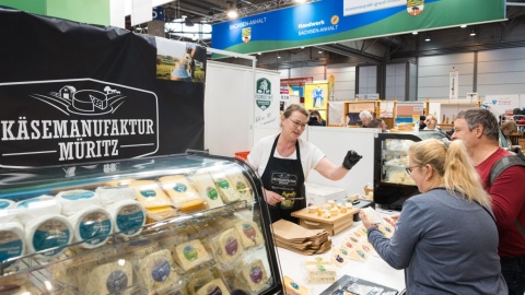 2 Personen lassen sich von einer Frau an einem Verkaufsstand für Käse beraten.