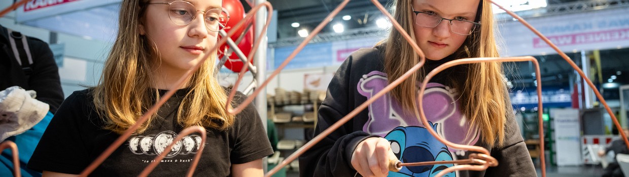 Zwei Mädchen experimentieren an einem Tisch mit gebogenen Kupferdrähten in einer Messehalle.