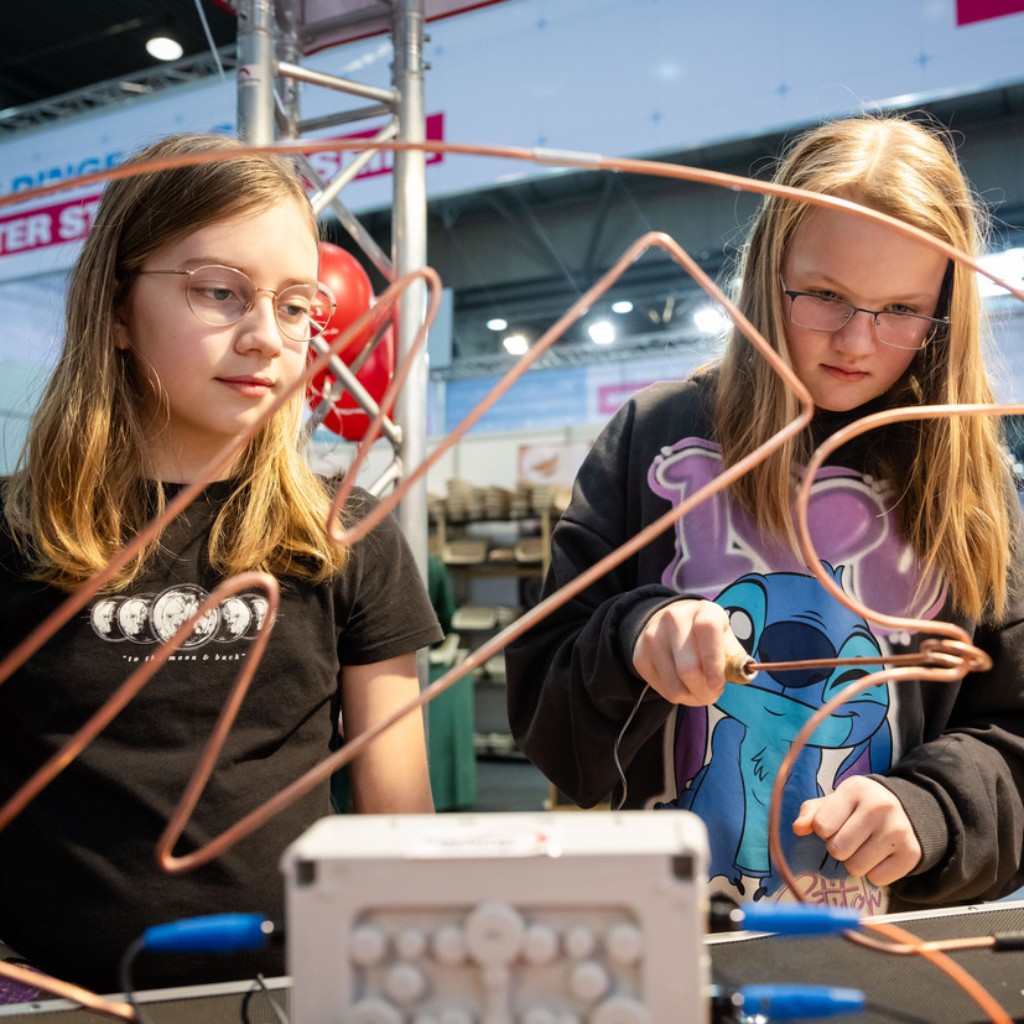 Zwei Mädchen experimentieren an einem Tisch mit gebogenen Kupferdrähten in einer Messehalle.