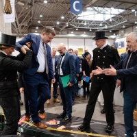 Ein Mann im Anzug balanciert auf einem Gurt und wird von 1 Person dabei festgehalten.